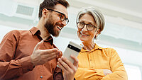Ein Mann und eine Frau schauen lächelnd gemeinsam auf ein Smartphone, was die Nutzung digitaler Gesundheitsanwendungen im Alltag symbolisiert.