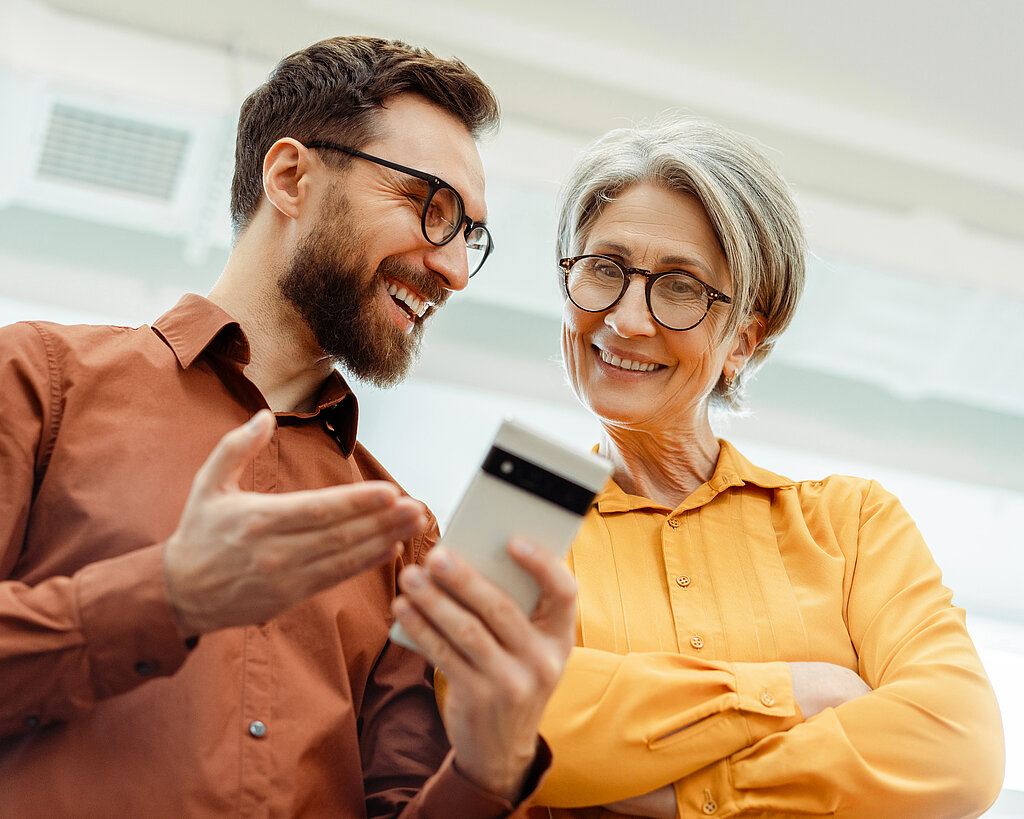 Ein Mann und eine Frau schauen lächelnd gemeinsam auf ein Smartphone, was die Nutzung digitaler Gesundheitsanwendungen im Alltag symbolisiert.