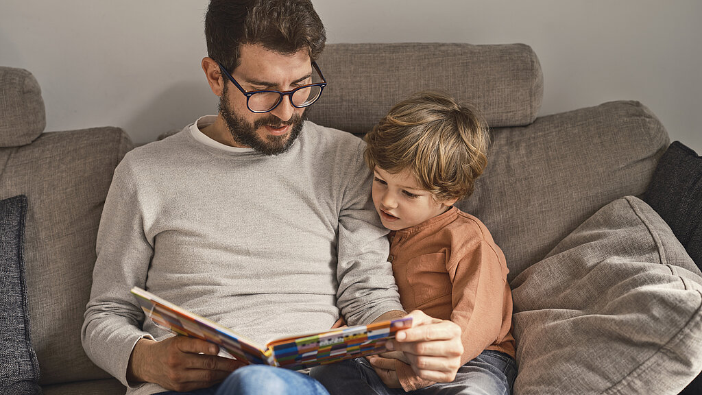 Ein Vater sitzt mit seinem  Sohn auf dem Sofa und liest aus einem Buch vor