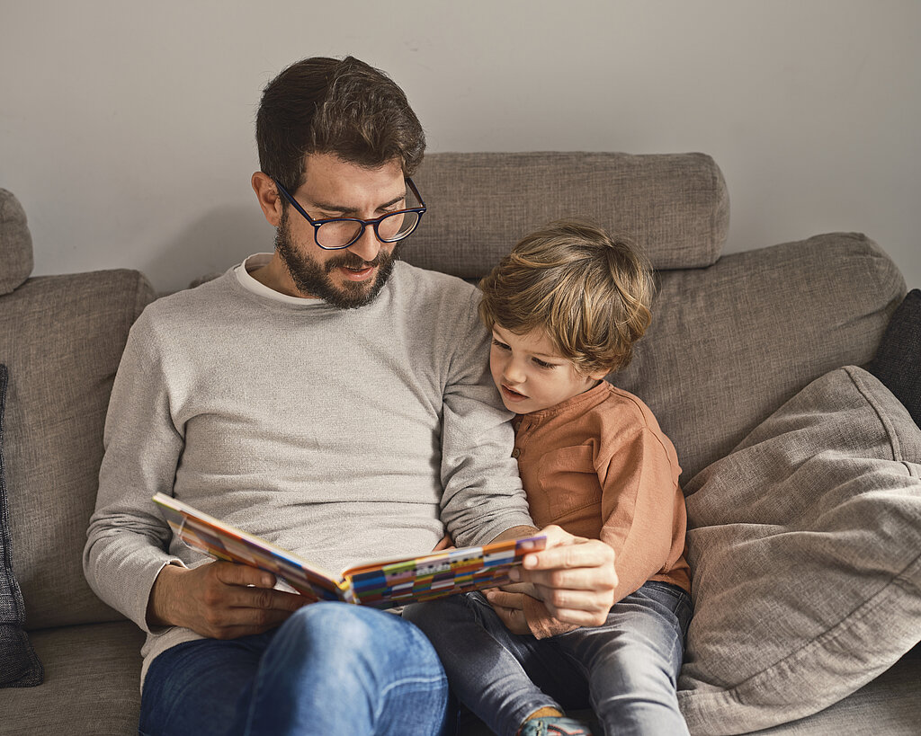 Ein Vater sitzt mit seinem  Sohn auf dem Sofa und liest aus einem Buch vor